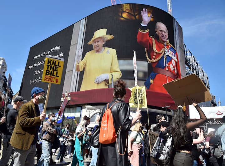 Protesters walk past images of Britains Prince Philip displayed on a giant screen at Piccadilly Circus, London. The funeral started at 2.40 p.m. (local time) with the coffin leaving the Castle, followed by a funeral march and a nationwide minute of silence, according to Buckingham Palace. (Image: PTI)