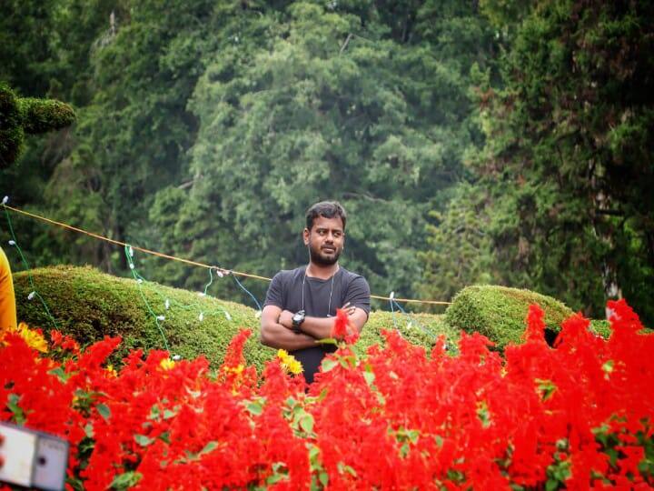 மலர்கண்காட்சிக்கு நடுவே இளைஞர் ஒருவர் புகைப்படம் எடுத்துக் கொள்ளும் காட்சி.