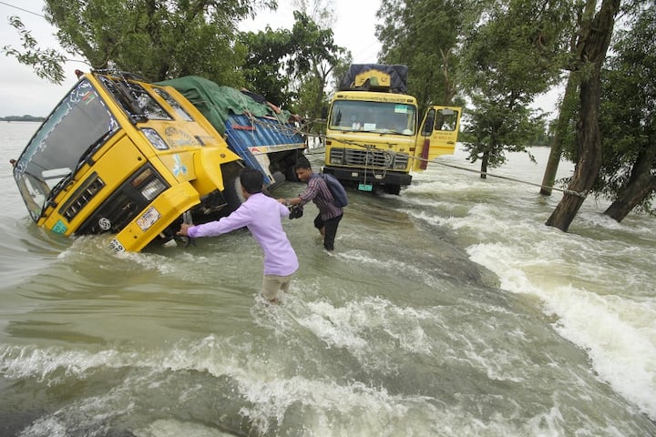 People wade past stranded trucks on a flooded street in Sunamganj on June 21. Massive floods have ravaged parts of Bangladesh, leaving dozens of people dead and millions stranded. Floods are a regular menace to millions of people in low-lying Bangladesh, but experts say climate change is increasing their frequency. (Image Source: AFP)