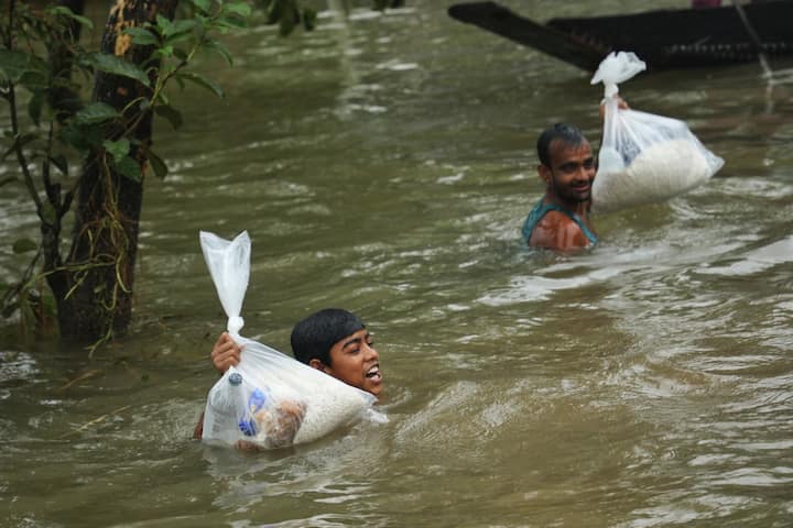 People wade through chest-deep flood waters after collecting food packets in a flooded residential area following heavy monsoon rainfalls in Companiganj. (Image Source: AFP)