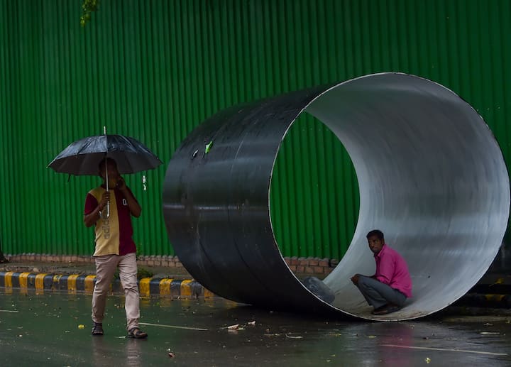 A man holding umbrella walks as another sits inside a pipe amid monsoon rains. (Image Source: PTI)