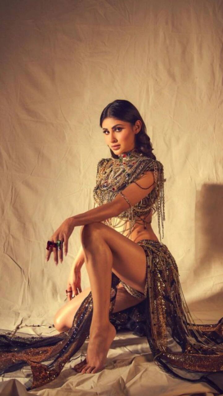 मौनी रॉयच्या नाट्यमय पोशाखात उंच गळ्याचा ब्लाउज, पूर्णपणे स्टोन आणि साखळ्यांनी जडलेला, कमी कंबर असलेल्या उंच स्लिट स्कर्टचा समावेश आहे.