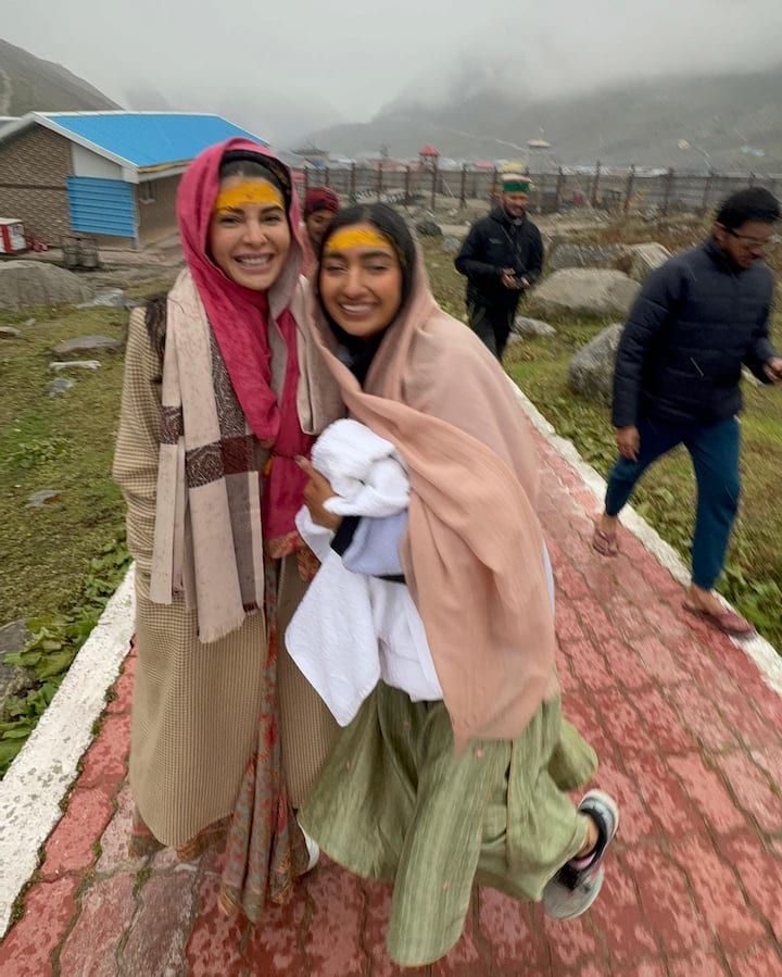 Jacqueline is wearing a multi-coloured Anarkali kurta, paired with a chequered trench coat, and a shawl. She has kept her hair open, with no makeup look, and white sneakers.