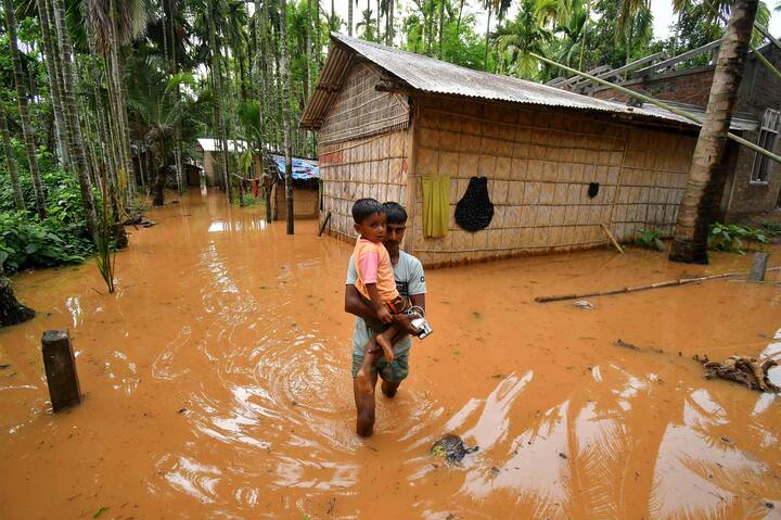 असम के नागांव जिले के कामपुर के पास सिंगी मारी में चक्रवात रेमल के आने के बाद भारी बारिश के बाद बाढ़ग्रस्त गांव में एक व्यक्ति एक बच्चे को गोद में लिए हुए है, बुधवार, 29 मई, 2024। (स्रोत: पीटीआई)