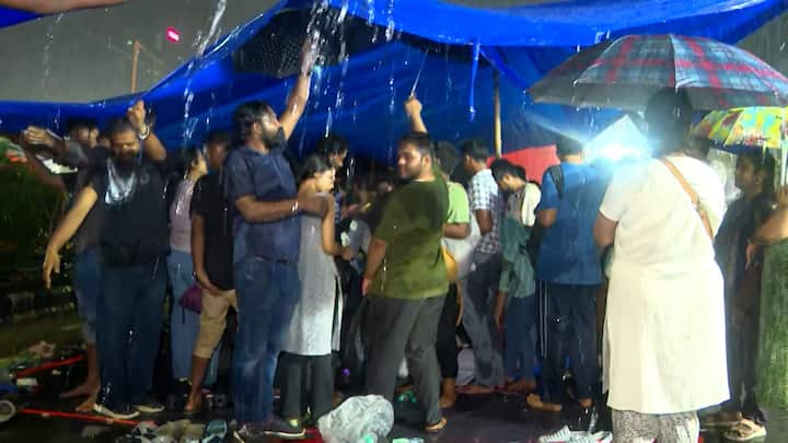 শুক্রবার দেখা গেল প্রবল বৃষ্টির মধ্যে ত্রিপল ধরে দাঁড়িয়ে রয়েছেন জুনিয়র চিকিৎসকরা। কেউ কেউ নিচে বসেও আছেন। তাঁদের শরীর ভিজে গেলেও কমেনি আন্দোলনের ঝাঁঝ।