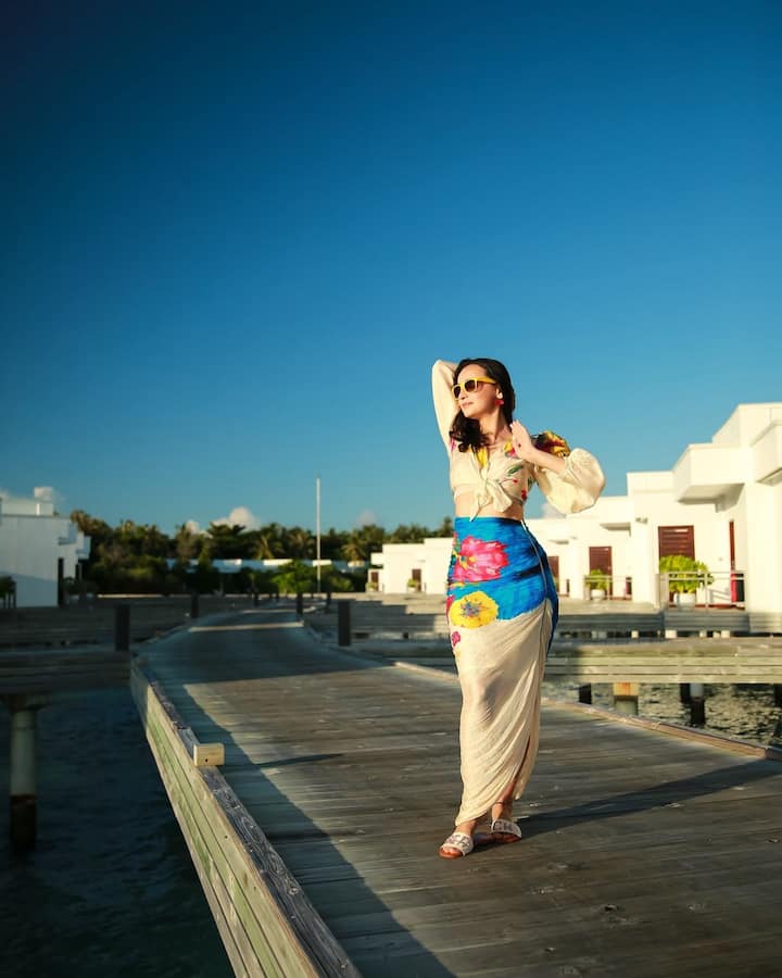 Complementing her look are stylish yellow sunglasses and earrings, rounding off the ensemble with breezy slippers that capture the essence of island chic.