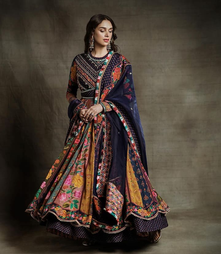 This beautiful multi-colored Anarkali suit features a three-piece churidar and a traditional odini, styled by Sanam Ratansi. The closed neck adds richness, while the hanging jhumka enhances its radiance, making it perfect for festive occasions like Durga Puja.