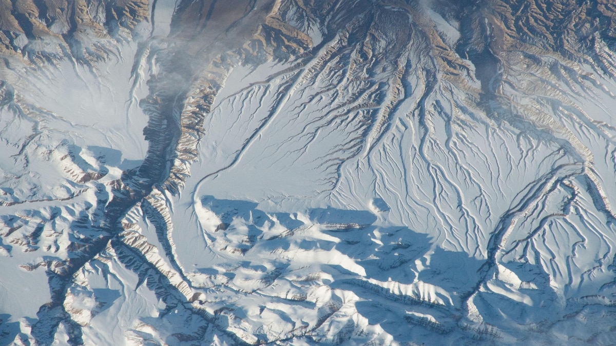 The Himalayas’ snowy peaks cast evening shadows in this oblique view. River erosion forms alluvial fans, with deep canyons slicing through the landscape, showcasing dramatic mountain topography.