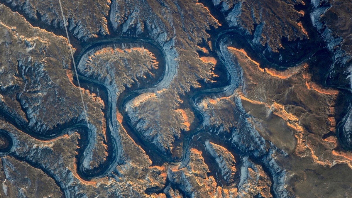 Bowknot Bend in Utah’s Green River canyon features striking meanders and steep cliffs. A jet contrail bisects the scene, revealing the interplay of natural and human activity.