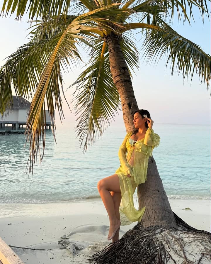 Neha leans casually against a palm tree, her confidence perfectly complementing the tranquil beach setting.