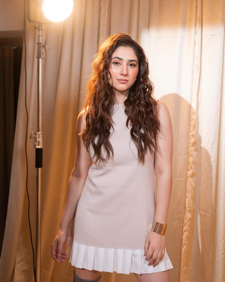 Disha Parmar commands attention in a creamy white mini dress, standing tall against a backdrop of golden drapes and studio lights.