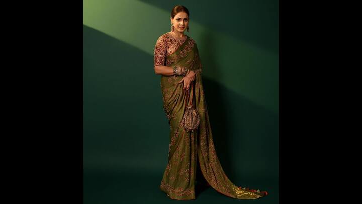 7. Bandhani Saree: Genelia D'Souza exuded classic elegance in an exquisite olive green bandhani saree. The saree consisted of tie and dye patterns and was finished with striking red border that added depth to the outfit. She paired her saree with a heavily embellished blouse that had mirror and threadwork in shades of maroon, rust, and gold. She wore chunky traditional bangles that added a festive spark to her fit along with statement earrings. (Image Source: Instagram/@geneliad)