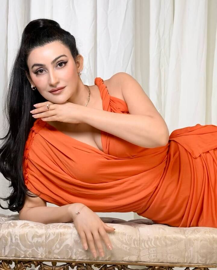 Shalini wears a striking orange draped gown, asymmetrically gathered at the shoulders with delicate pleats and structured tucks. The flowing silhouette is minimal yet impactful, allowing the vivid hue to take center stage.