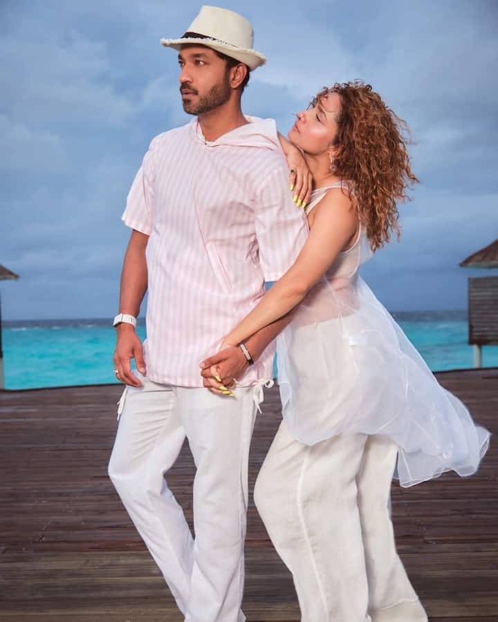 Captured on a wooden deck near a picturesque cottage, the duo poses confidently against the backdrop of the turquoise sea.