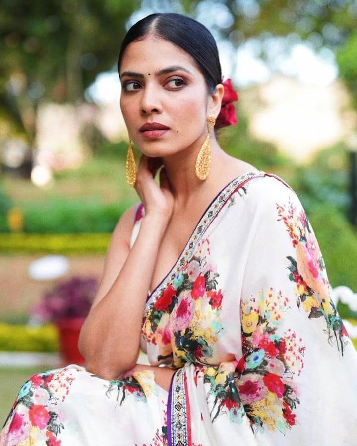 Draped in Floral Satin: She looked fresh and vintage-chic in a white silk satin saree with vibrant floral prints. The unique border, matching blouse, bold gold danglers, and rose-adorned bun gave this look a perfect retro touch. (All Image: Malavika Mohanan/Instagram.)