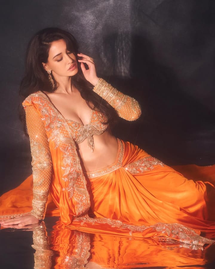 In this look, direction looks traditional and glamorous. He is wearing an orange and golden detailing ethnic outfit, giving a very royal and festive look. Open hair and minimal makeup with deep neck blouse and heavy embroidery are increasing their natural beauty.