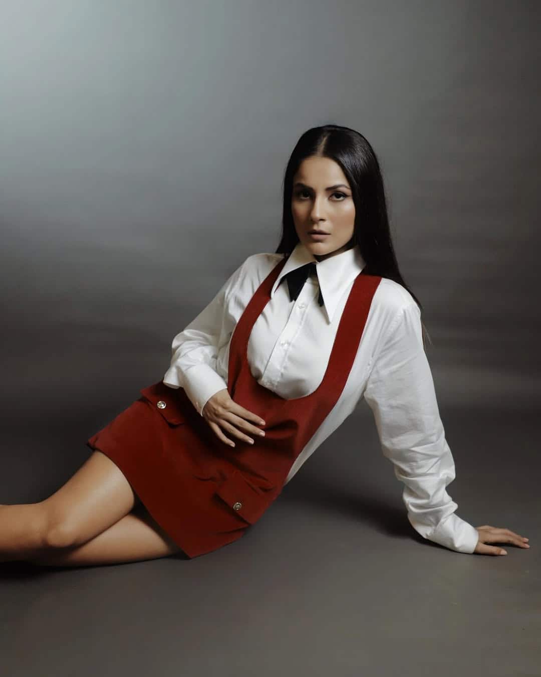 In this look, Shahnaz is wearing a maroon dress with a white shirt, which is giving her a smart look. The shirt's collar and the simple design of the dress is making this look softcharged. Straight is completing hair and nude makeup look.