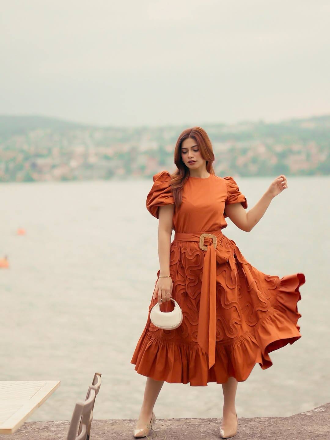 In this look, Nagma is wearing a rust orange dress which has belt detailing with puff sleeves and flared skirts. There are textured designs on the dress. They have pair it with nude heels and small white handbags. Open hair and satal makeup is giving this look modern touch.