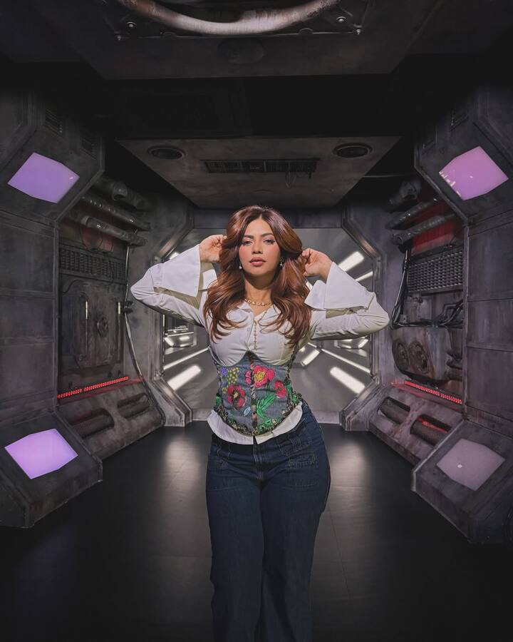 In this look, Nagma is wearing a white colored bell-sleeve top, which is paired with a corset belt with colorful flowers, giving a touch in her look. He has pair this outfit with dark blue jeans. Open and light waves, light makeup and nude lipstick are completing her look.