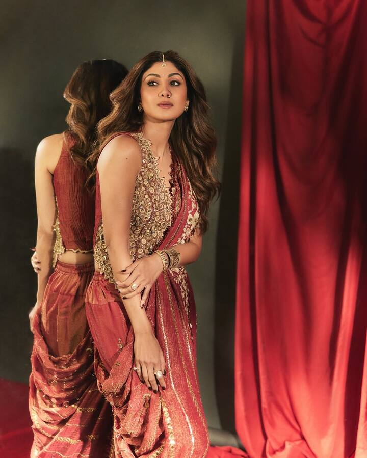 इस लुक में शिल्पा बेहद ट्रेडिशनल अंदाज़ मे दिखाई दे रहा है. उन्होंने डार्क रेड रंग का शानदार ड्रेप-स्टाइल आउटफिट पहना है, जिस पर गोल्डन कढ़ाई और डिटेलिंग की गई है. उनके ब्लाउज़ पर किए गए बारीक काम और दुपट्टे जैसी पल्लू स्टाइलिंग इस ड्रेस को थोड़ा अलग लुक दे रही है. खुले, वेवी बाल और हल्के ग्लैम मेकअप के साथ माथे पर छोटा सा मांगटीका उनके लुक में ट्रेडिशनल चार्म ऐड कर रहा है. हाथों में चूड़ियां और अंगूठियां उनके इंडियन एथनिक लुक को पूरा कर रही हैं.