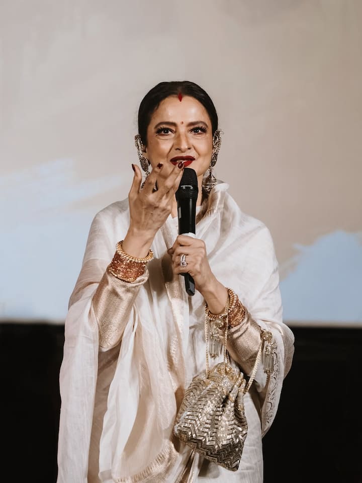 इस लुक में रेखा ने सफेद सिल्क साड़ी पहनी है, जिसे गोल्डन बॉर्डर ने और खास बना दिया है. कानों में स्टेटमेंट ज्वेलरी, लाल बिंदी और लाल लिपस्टिक उनके लुक को क्लासिक टच देती है. हाथ में गोल्डन पॉटली बैग उनके पूरे अंदाज़ को एलीगेंट और ग्रेसफुल बना रहा है.