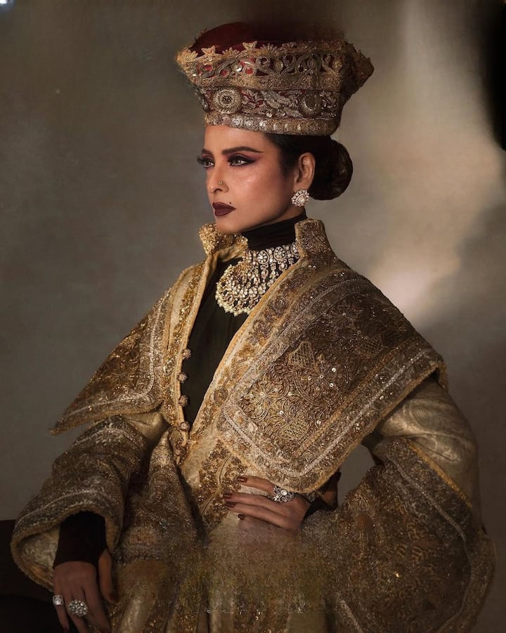 रेखा ने इस लुक में गोल्डन और भारी कढ़ाई वाले आउटफिट के साथ जड़ाऊ गहने पहने हैं. सिर पर बड़ा हेडगियर उन्हें महारानी जैसी लुक दे रहा है. गहरे आई-मेकअप और डार्क लिपस्टिक के साथ उनका यह लुक एलीगेंस को बखूबी दिखा रहा है.