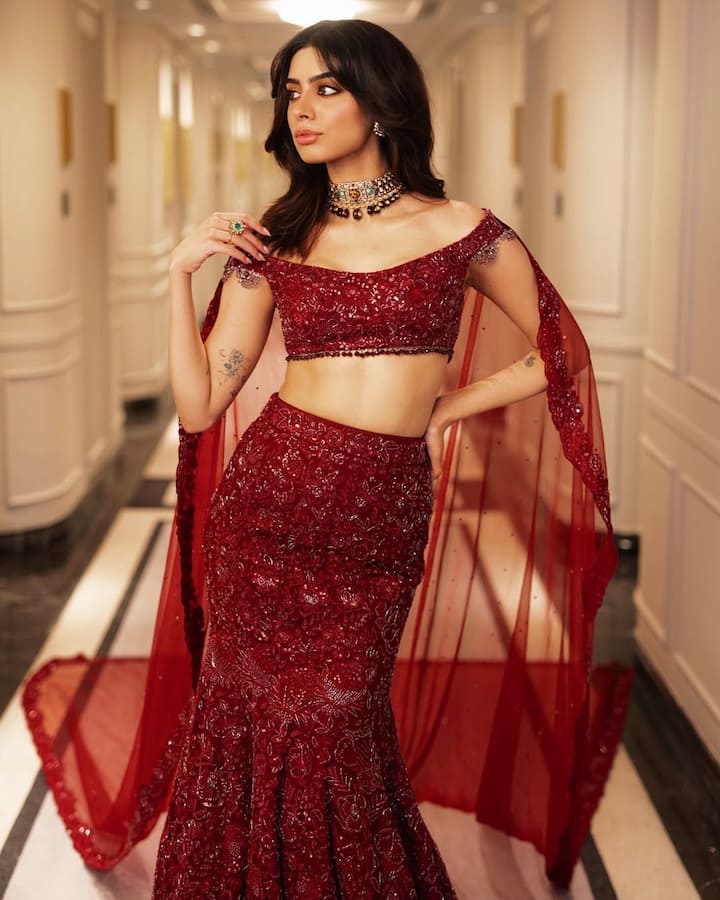 This look of happiness is very glamorous and stylish. He is wearing a dark red-colored heavy embroidery lehenga, with an off-shoulder blouse and a net scarf. The statement around the neck is giving a royal touch to his look. Open wavy hairstyle and minimal makeup are further enhancing their beauty.