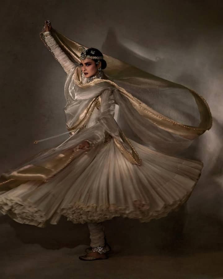 रेखा इस लुक में एक दम मुग़ल-ए-आज़म की अनारकली जैसी लग रही हैं — रॉयल, ग्रेसफुल और टाइमलेस. वो एक घेर वाला अनारकली सूट पहने हुए हैं, जो गोल्डन बॉर्डर के साथ रॉयल ऑफ-व्हाइट रंग में है. सिर पर माथापट्टी और भारी जड़ाऊ गहने उनकी खूबसूरती को और निखार रहे हैं. इस तस्वीर में वो कथक स्टाइल में घूमती हुई नज़र आ रही हैं, जिससे उनका लुक और दुपट्टा बेहद खूबसूरती से लहरा रहा है.