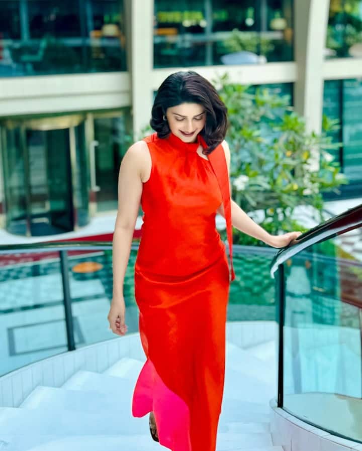 This look of Prachi Desai is very classy. She is wearing a sleeveless red-orange high neck gown with a slight hot pink shed at the bottom, which gives the outfit fusion and fun touch. The straight fall of the gown and the satisfied finish, the stylish bo tie tied on the high neck adds the drama to the detail look. The hairstyle is in soft waves, which makes the look more feminine and somber. Makeup and accessories are minimal so that the whole focus goes to the outfit.
