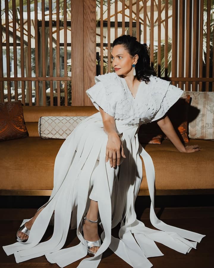 Maria is wearing a white drape-style outfit in this look. Silver heels and statement earrings are making their look even more glamorous. His look looks modern with sleek hairstyle and soft makeup.