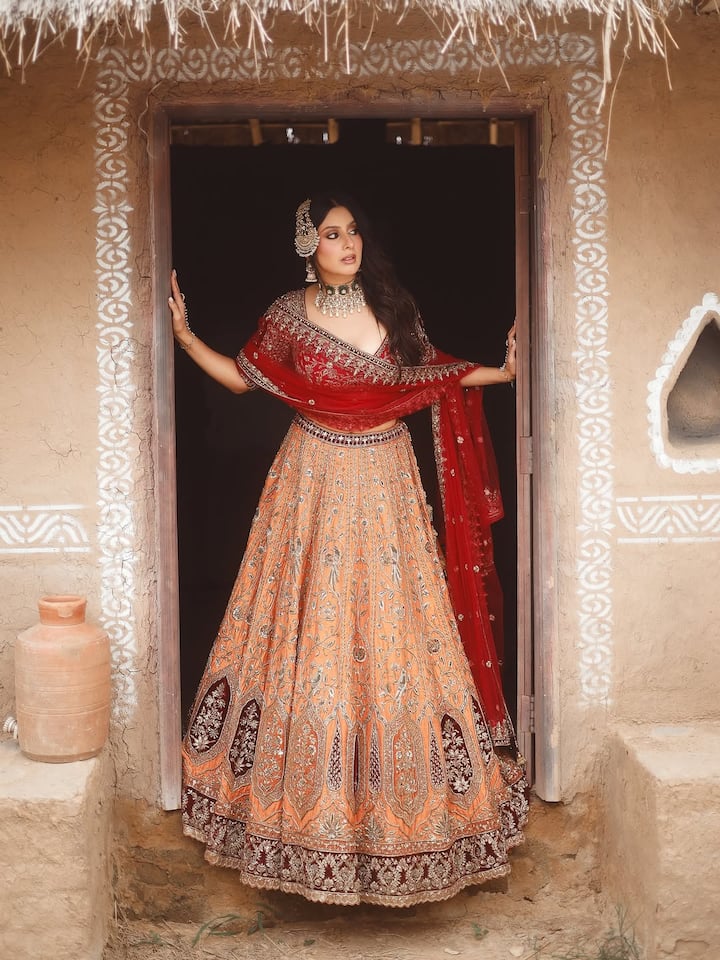 इस लुक में ईशा ने ट्रेडिशनल और रॉयल अंदाज़ अपनाया है. उन्होंने ऑरेंज और मरून शेड का हैवी एम्ब्रॉइडर्ड लहंगा पहना है, जिसे रेड दुपट्टे और डीप नेक ब्लाउज़ के साथ स्टाइल किया है. हैवी ज्वेलरी, चोकर नेकलेस और माथा-पट्टी उनके लुक को और भी रॉयल बना रही हैं. मिट्टी के घर और देसी बैकड्रॉप में उनका ये ट्रेडिशनल अवतार बेहद एथनिक वाइब्स दे रहा है.