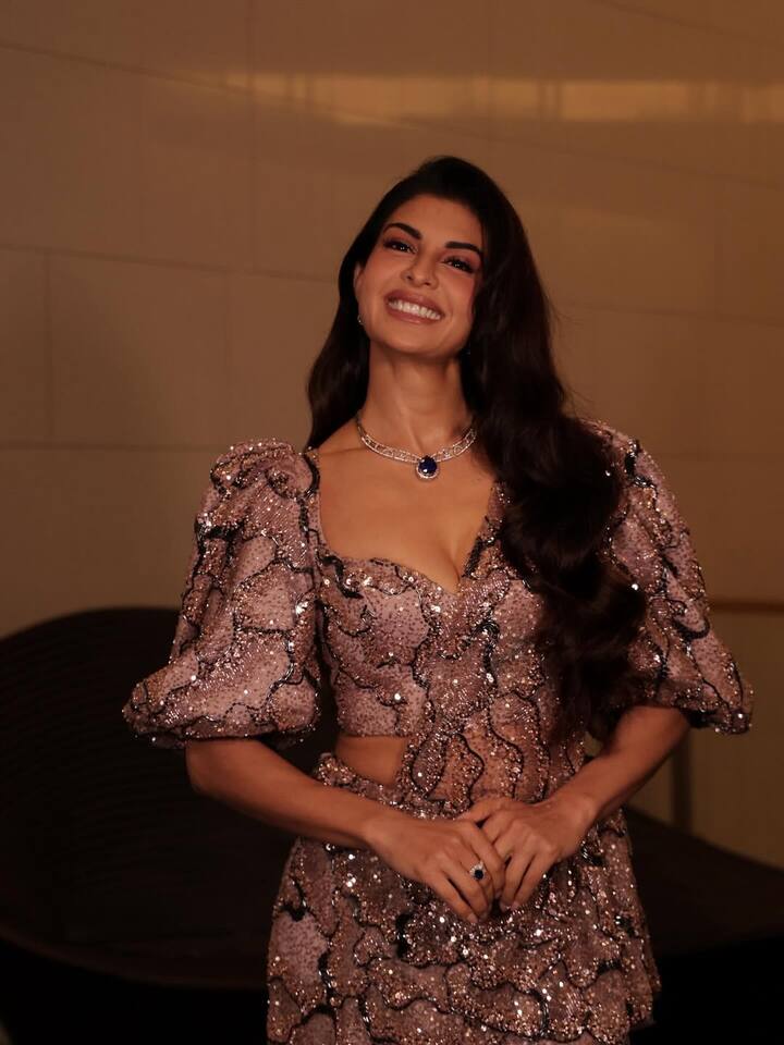 In this look, Jacqueline is wearing a lavender-pink outfit with Shimri sequin, which has puffy sleeves and stylish cut-out design. The elegant necklace of open wavy hair, nude makeup and blue stone is giving her look even more Royal Touch.