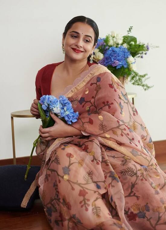 इस तस्वीर में विद्या बालन फ्लोरल प्रिंटेड साड़ी और रेड ब्लाउज में बेहद प्यारी लग रही हैं. हाथों में नीले फूल और मुस्कान उनके लुक को और भी खास बना रही है.