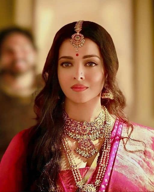 इस तस्वीर में ऐश्वर्या राय बच्चन बेहद रॉयल और ग्रेसफुल अंदाज में नजर आ रही हैं. उन्होंने गुलाबी रंग की साड़ी पहनी है, जिस पर गोल्डन काम है. उनके लुक को और निखार रहा है भारी गोल्ड ज्वेलरी सेट, जिसमें लंबी हार, चोकर, झुमके और माथे पर सुन्दर मांगटीका शामिल है. खुले बाल और हल्का मेकअप उनके चेहरे की खूबसूरती को और भी उभार रहा है.