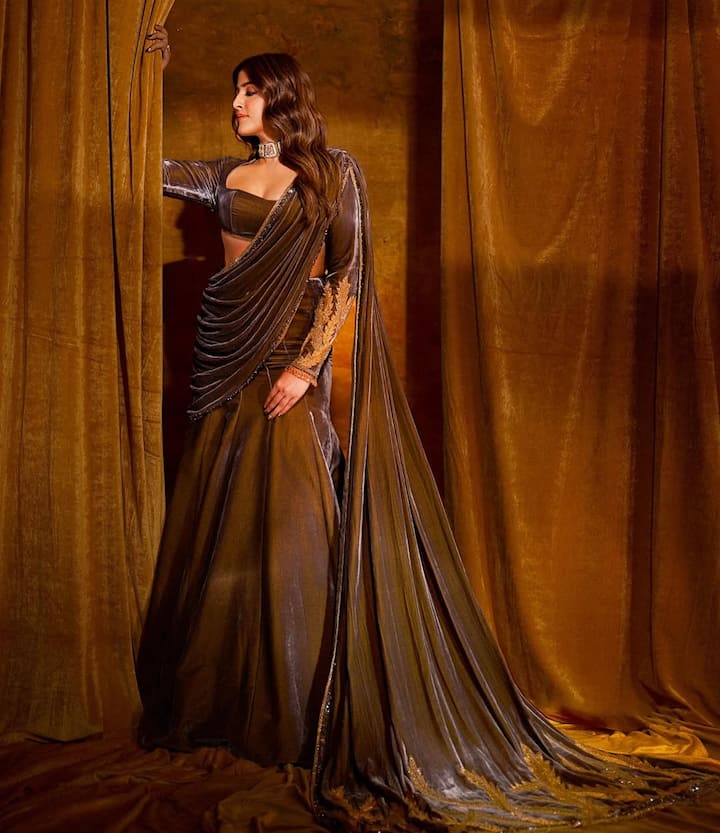 Nupur is wearing a very stylish and modern saree. This saree is gray-gold colored and is made of velvet cloth. She is wearing this saree with a corset-style blouse. The pallu of the saree is very long and has been dragged from one side.