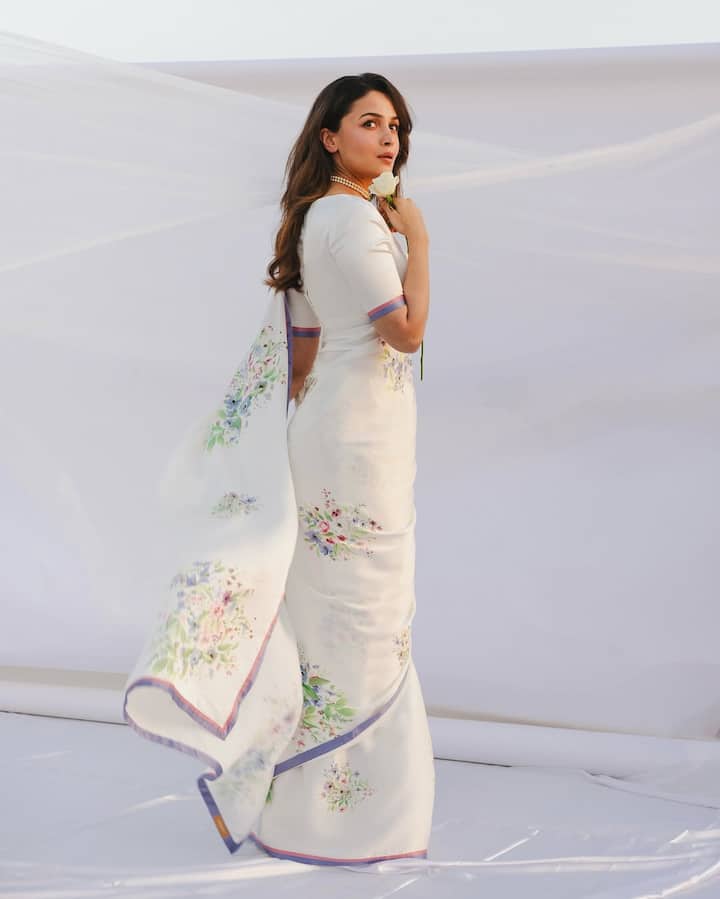 Alia is wearing a beautiful white colored saree in this look. She has paired this saree with a sum, white blouse. The design of the blouse is very simple, which has sleeves up to round throat and albow. On the edge of the sleeves, there is a blue and pink bandage matching with the border of the saree. This blouse is an excellent exam of designs and elegance.