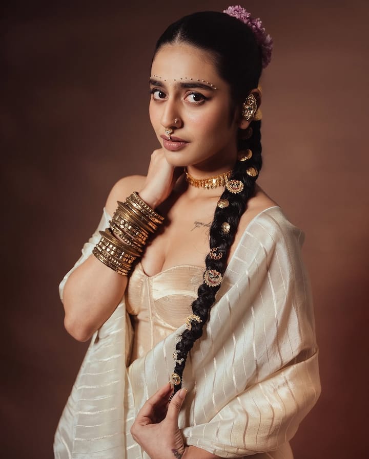 Priya has draped off white saree in separate style with a crosset blouse here. Make a braid with golden jewelery and applying flowers to the hair, she is looking very classy.