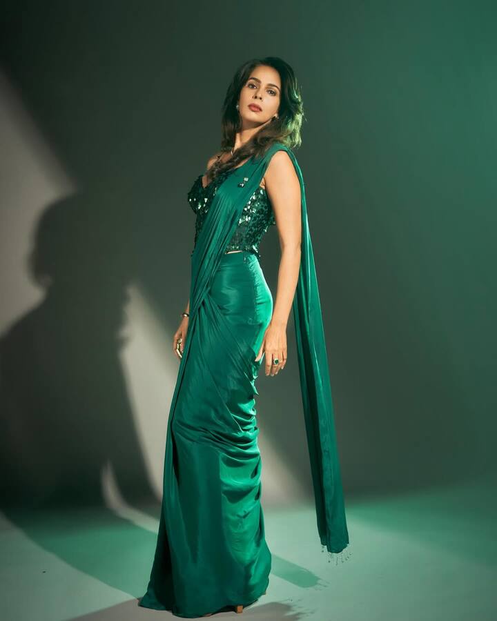 In this look, Mallika is wearing dark green satin saree. She has pairing the saree with a sleeveless blouse with matching sequences, giving her a modern party look. They have kept the accessories low, wearing only a few rings and thin bracelets, causing the color of the saree and highlight. She has kept the hair open and standing in the side pose, she is showing her figure which is making this look quite catch.