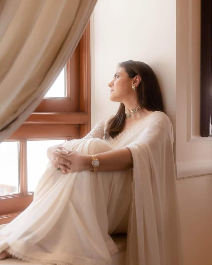 Kajol is in a beautiful, off-white colored saree for this look. She is wearing this saree with a matching full-salivary blouse, which has a light embroidery. In an accessory, he is wearing a gold bran necklace with green pearls, and also wearing matching earrings. He is wearing a clock in one hand and a ring in one. Their hair is open and makeup is very light. This entire look looks very somber and elegant.