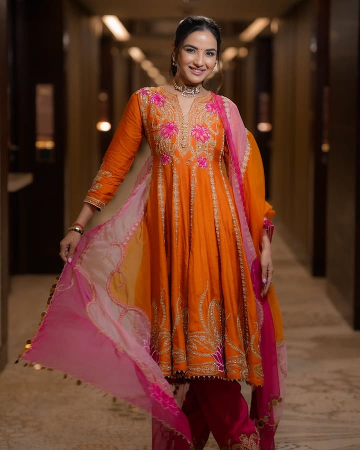 इस लुक में जैस्मीन नारंगी रंग की एक छोटी अनारकली-स्टाइल कुर्ती में हैं. कुर्ती पर गुलाबी और गोल्डन धागों से बारीक कढ़ाई की गई है, खासकर गले और बीच के हिस्से में, जो एक सुंदर पैटर्न बना रहा है. उन्होंने नीचे गुलाबी रंग की पटियाला स्टाइल पैंट पहनी है. दुपट्टा गुलाबी और नारंगी रंग का शेडेड है, जिसके किनारों पर गोटे का काम है. एक्सेसरीज़ के लिए उन्होंने चोकर नेकलेस और हाथ में कंगन पहने हैं. उनका यह पूरा लुक फेस्टिव वाला लग रहा है.