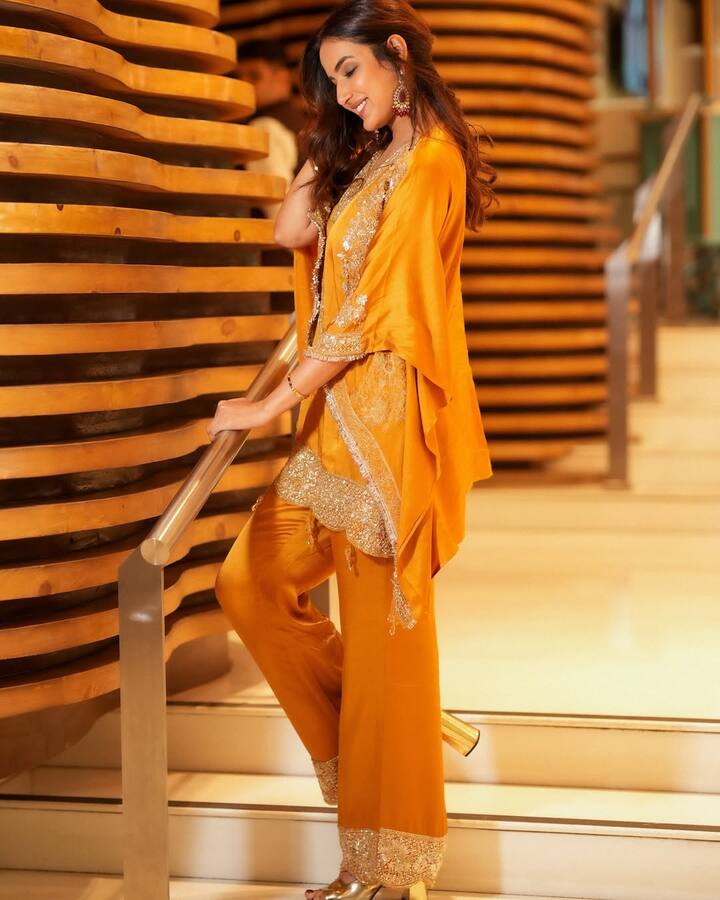 इस फोटो में जैस्मीन ने पीले रंग का एक एथनिक सेट पहना है. यह एक काफ्तान-स्टाइल का कुर्ता है जिस पर बारीक गोल्डन एम्ब्रॉइडरी का काम किया गया है. उन्होंने इसे चौड़े, पलाज़ो-स्टाइल के पैंट के साथ पहना है, जिसके निचले हिस्से पर भी गोल्डन काम की पट्टी है. उनके पैरों में गोल्डन रंग की ऊँची हील्स हैं और उन्होंने बड़े गोल्डन झुमके पहने हैं. उनका मेकअप नेचुरल है और बाल खुले हैं, जो उनके फेस्टिव लुक को पूरा कर रहा है.