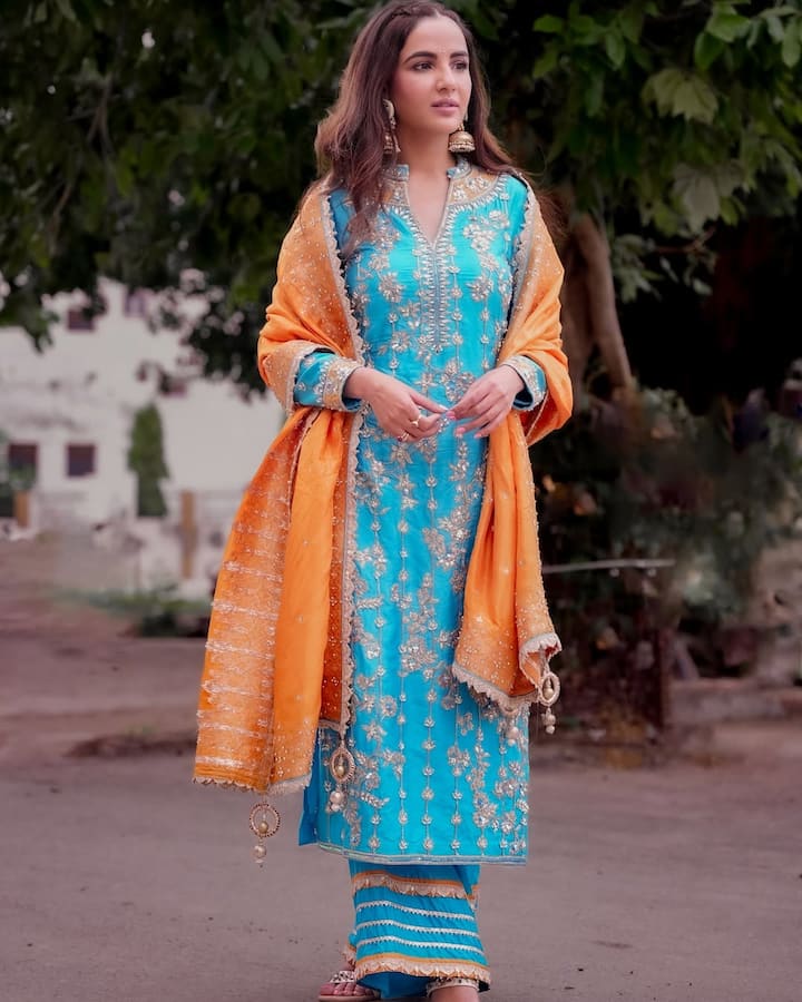 इस लुक में, जैस्मीन ने नीले रंग का एक पंजाबी स्टाइल का सलवार-सूट पहना है. कुर्ते और पैंट दोनों पर बारीक गोल्डन धागे की  एम्ब्रॉइडरी का काम है, जिसमें फूल-पत्तियों का डिज़ाइन दिख रहा है. उन्होंने इसे नारंगी रंग के दुपट्टे के साथ पेयर किया है, जिस पर भी किनारों पर गोल्डन टैसल्स लगे हैं, जो एक शानदार कलर कंट्रास्ट दे रहा है. उन्होंने अपने बाल खुले रखे हैं और बड़े, ट्रेडिशनल गोल्डन झुमके पहने हैं.