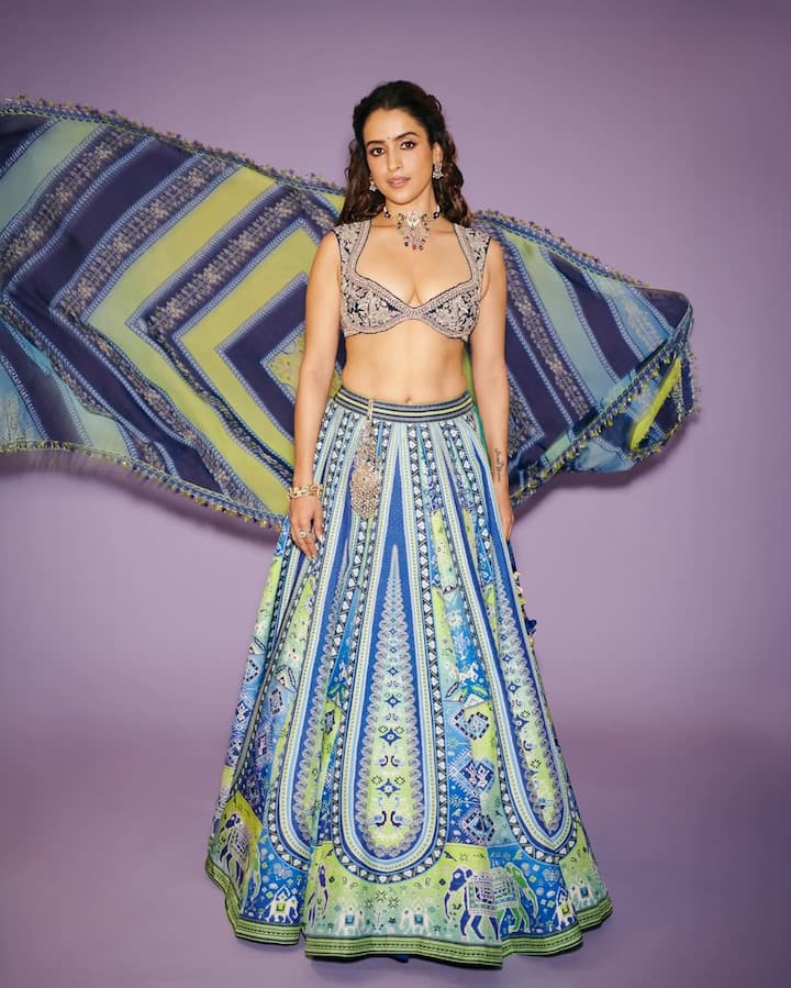 This look is a fresh and colored choice for the festive season. In this look, Sanya is wearing a beautiful digital printed lehenga with a print of blue and lime green colors, on which traditional motifs are made. He has paired the lehenga with a brulette styling choli with deep-neck, black and silver embroidery. Floi dupatta matching the lehenga is adding drama to this look. The accessories have oxidized silver bran and a waistband, giving the entire outfit a boho-ethnic vibe. This look is especially perfect for functions like turmeric, mehndi or garba-dandia.