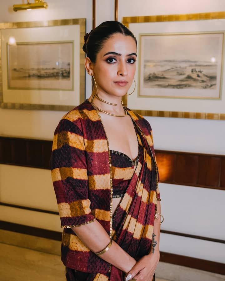 This look of Sanya is a traditional but trendy and different style for the festive season. She is wearing a modern saree of maroon, yellow and brown strip patterns, paired with a checked checked jacket. This jacket is giving an Indo-Western layered look to the saree, which is also good for the festivals of cold weather. He is wearing a strapy, deep-cut blouse. For accessories, there are golden bran-necklaces and large hops, giving the look a contemporary touch. This outfit is best for those who want to look different and stylish in festivals.