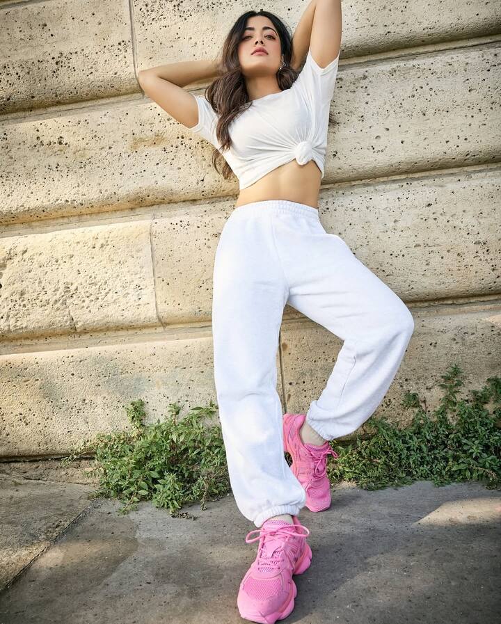 Rashmika is in a very cool and athletic look in this picture. He is wore completely white colored casual clothes, with high-waste joggers and a white crop T-shirt above, which is styled by tie a knot from the front. This entire white look is making a luxurious and trendy contrast with their bright pink sneakers. With open hair and a relax pose, this look is perfect for comfortable and stylish summer fashion.