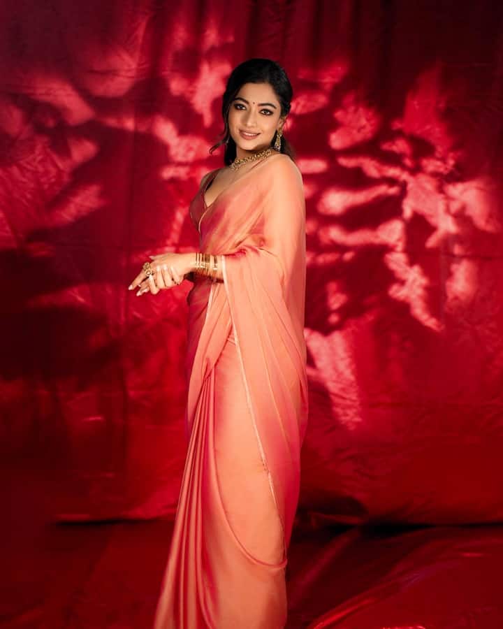 Rashmika is seen in a beautiful and elegant peach -colored plane chiffon saree in this picture. This saree is completely plain, with a thin golden border on the edges. He is wearing it with a deep-neck matching blouse. In an accessory, she is wearing a golden choker necklace, matching earrings and many golden bangles in her hands. Their hair is tied back, and their makeup is Satal, with a cute smile on his face, giving him a festive and graceful look.