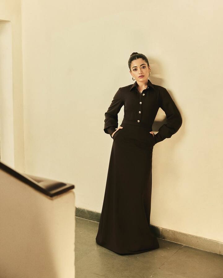 In this look, Rashmika is wearing a chocolate brown maxi dress, giving a shirt -like look, which has full sleeves and collar. This dress is going down like an A-line skirt, giving them a long and sleek appearance. Rashmika has tied her hair in a neet bun and wearing small hop earrings, causing her minimal and sophisticated look to complete. She is giving a confident pose by placing her hand on her waist.