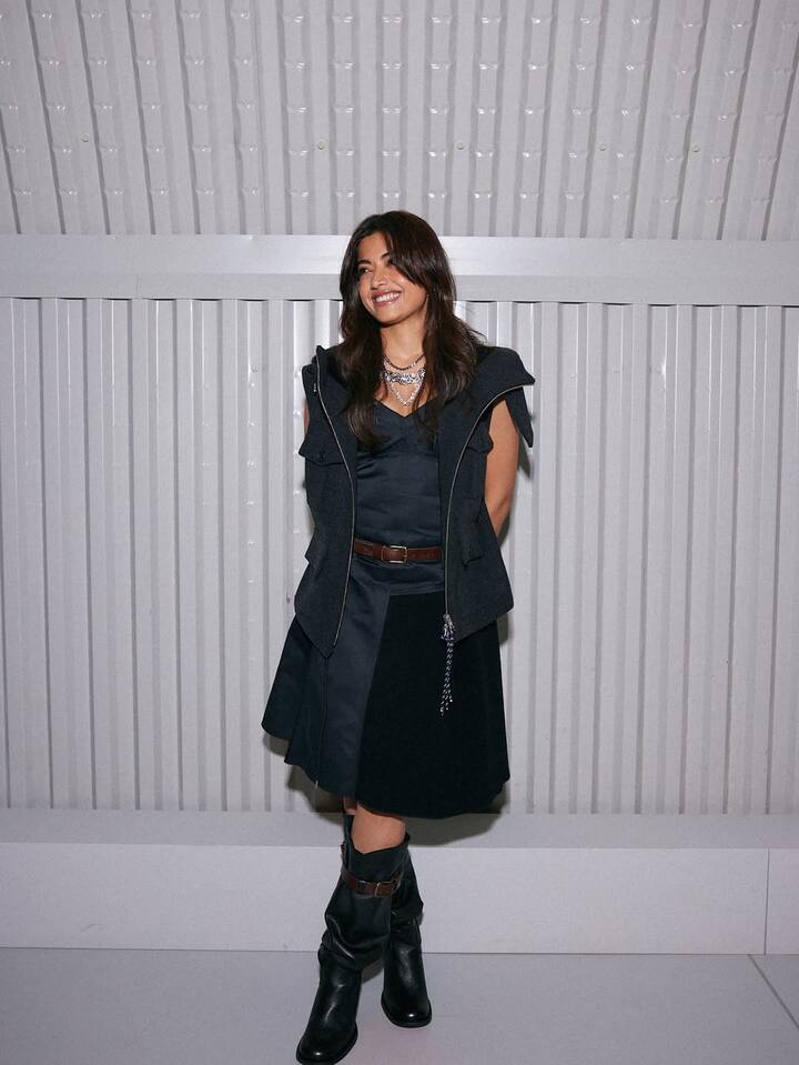 In this picture, Rashmika has carried a dark, utility-intelligent look. He is wearing a black nee-length dress, with a sleeveless waste layer, which has pockets and zipper detailing. There is a thin belt on the West, highlighting the Westline. He has completed this look with high black boots and a thick silver chain neckpiece. His look with open, medium length hair and a cute smile is rough-end-tuff and fashionable.