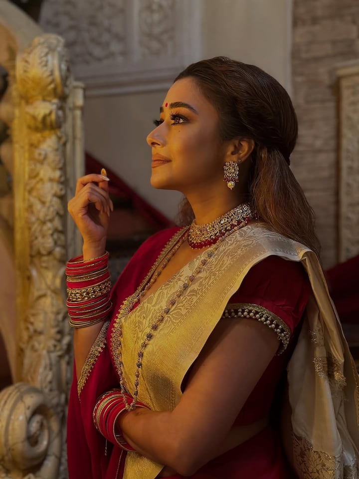 The saree’s fabric glistens softly under the golden lights, while the intricately woven borders with zari detailing add regal refinement. The red blouse, featuring delicate pleats on the sleeves, gives a structured, old-world charm that balances poise with contemporary sophistication.