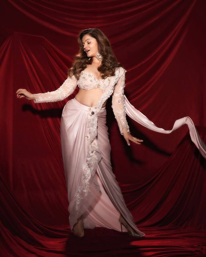 रुबीना ने यहां एक सॉफ्ट पिंक रंग की प्री-ड्रेप्ड शिमरी साड़ी पहनी है, जो करवा चौथ के लिए बेहद खूबसूरत लुक दे रही है. उन्होंने साड़ी को फुल-स्लीव वाले बारीक एम्ब्रॉयडरी वाले कॉर्सेट स्टाइल ब्लाउज के साथ पेयर किया है. साड़ी पर फ्लोरल डिटेलिंग इसे और भी फेमिनिन बना रही है. कर्ली बाल और हैवी झुमके उनके लुक को पूरा कर रहे हैं.